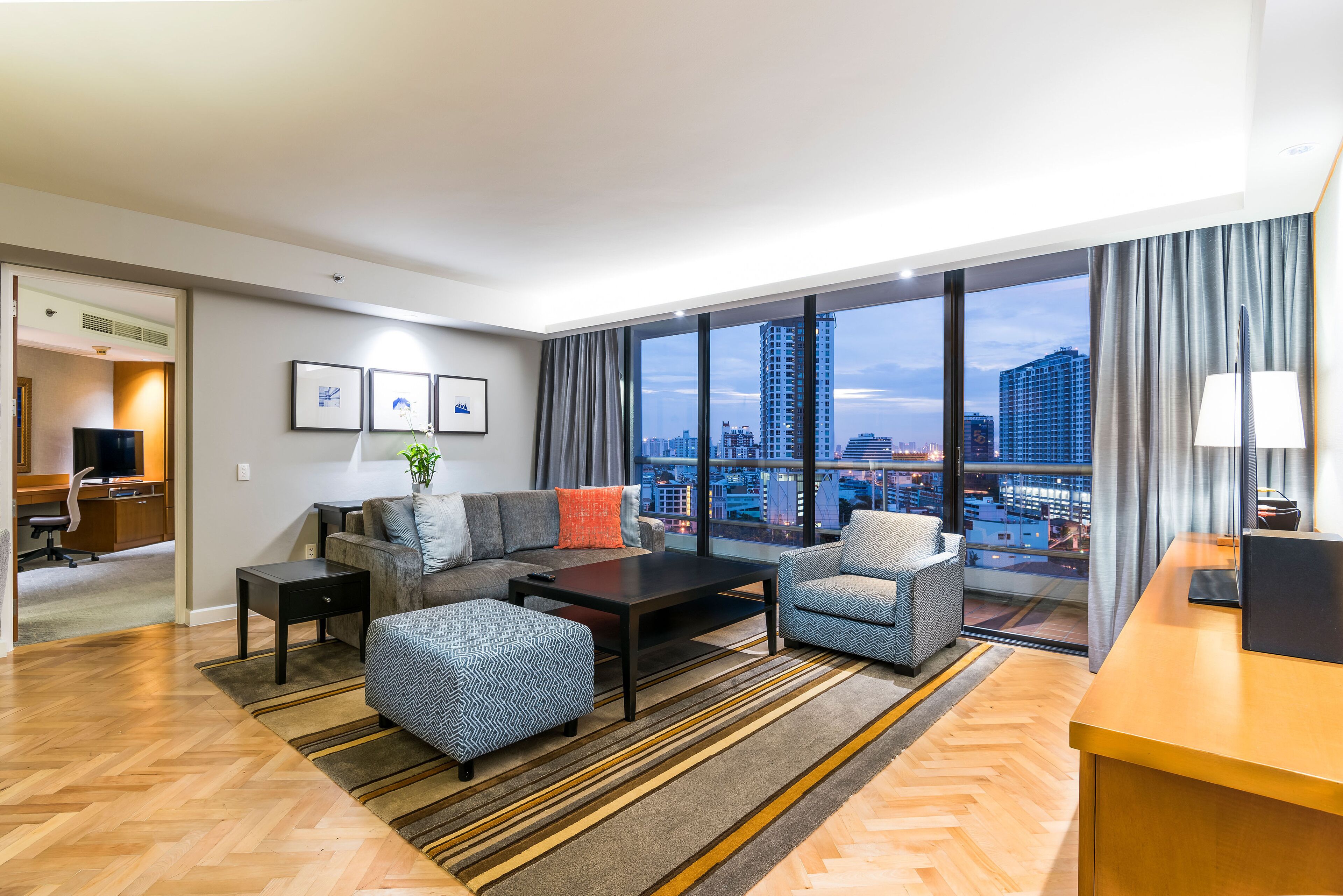 A modern hotel room with a large window offering a city view, a sofa, a coffee table, and a desk area.