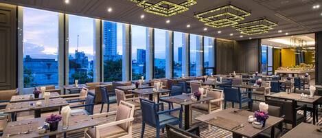 A spacious restaurant with multiple dining tables and chairs, large windows offering a city view, and modern geometric ceiling lights.