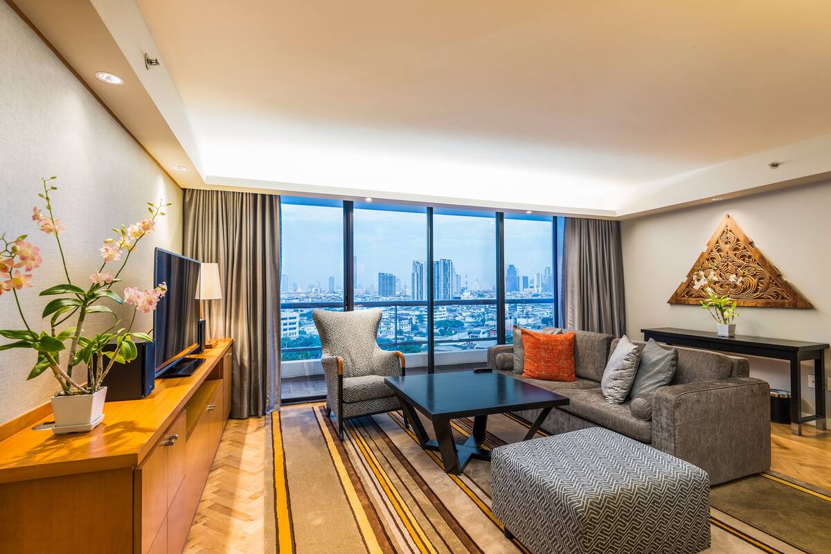 Ein modernes Wohnzimmer mit einer Couch, Hocker und Couchtisch. Ein Fernseher steht auf einem Schrank, eine Zimmerpflanze und Blick auf die Stadt durch große Fenster.