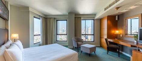 A hotel room with a large bed, a desk, a chair, and a TV. There are two windows with city views.