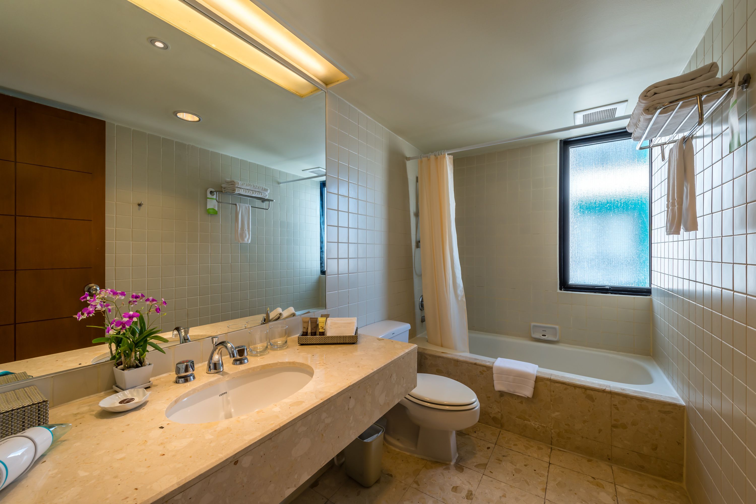 A modern bathroom with a large mirror, dual sinks, a toilet, a bathtub, and a window.
