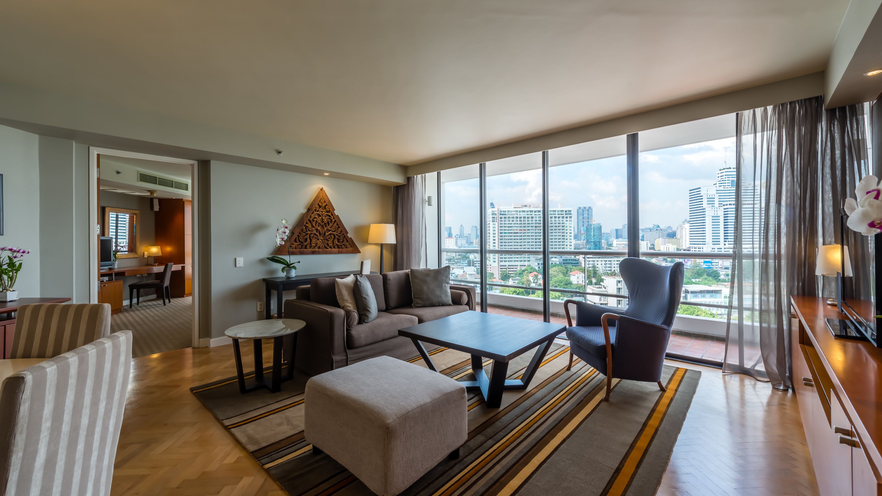 A modern hotel room with a large window offering a city view, a cozy seating area with a sofa and ottoman, and a small desk with a chair.