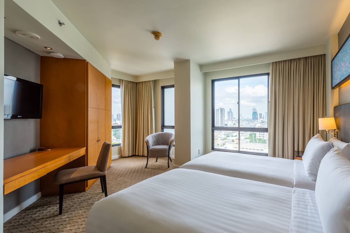 Ein Hotelzimmer mit zwei Betten, einem Schreibtisch, einem Sessel, einem Fernseher und Blick auf die Stadt.