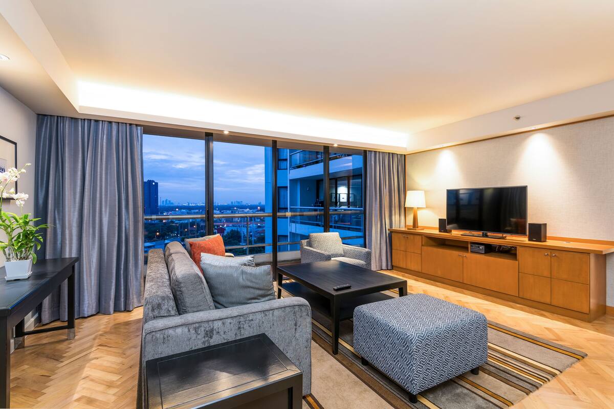 Ein modernes Hotelzimmer mit großem Fenster und Blick auf die Stadt, einer Couch, einem Couchtisch, einem Fernseher und einem Holzkasten.