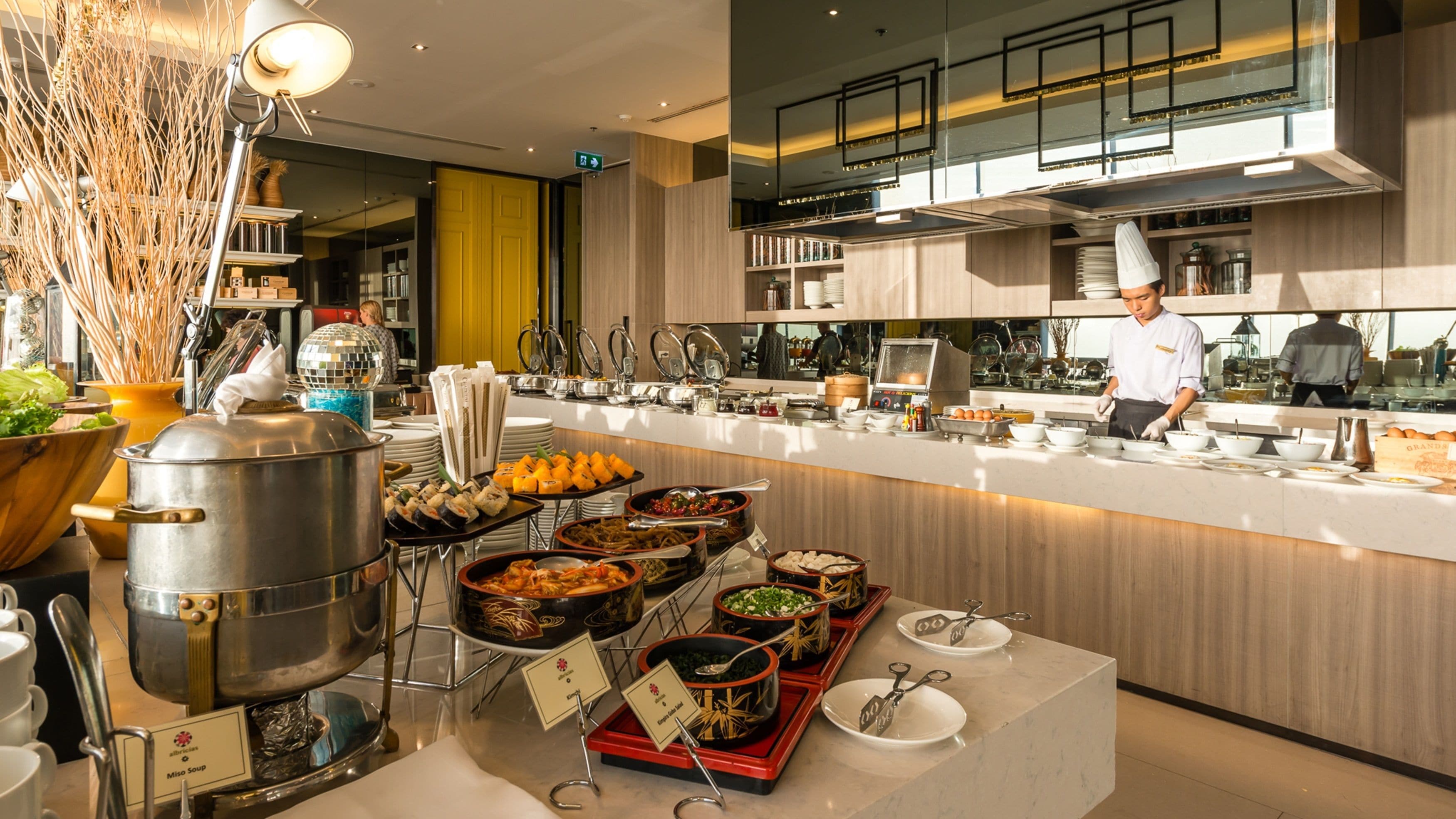 Buffet com diversos pontos de comida, um chef preparando alimentos e uma cozinha moderna ao fundo.