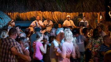 Uma banda se apresentando em um palco com um painel de cobertura de palha, cercada por uma multidão.