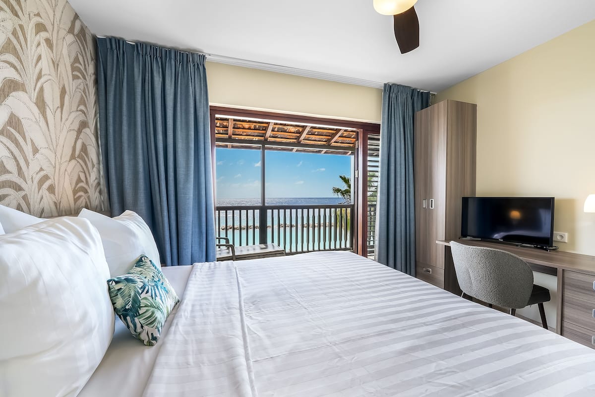 A hotel room with a bed, a desk, a television, and a balcony with a view of the sea.