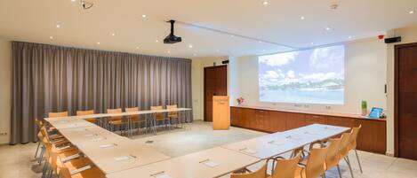 A conference room with a long table, chairs, a podium, a projector screen, and a window with a view.