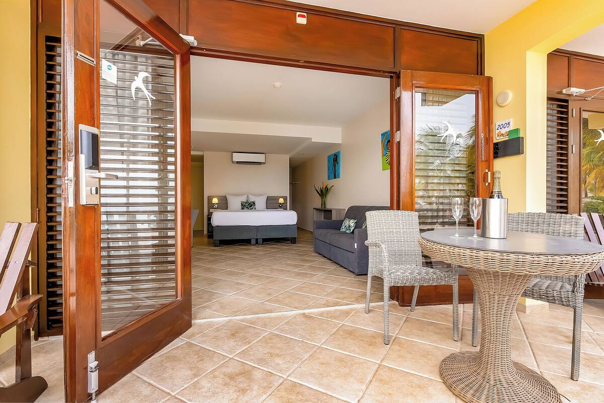 A hotel room with a bed, a sofa, a dining table with chairs, and a balcony with a view.