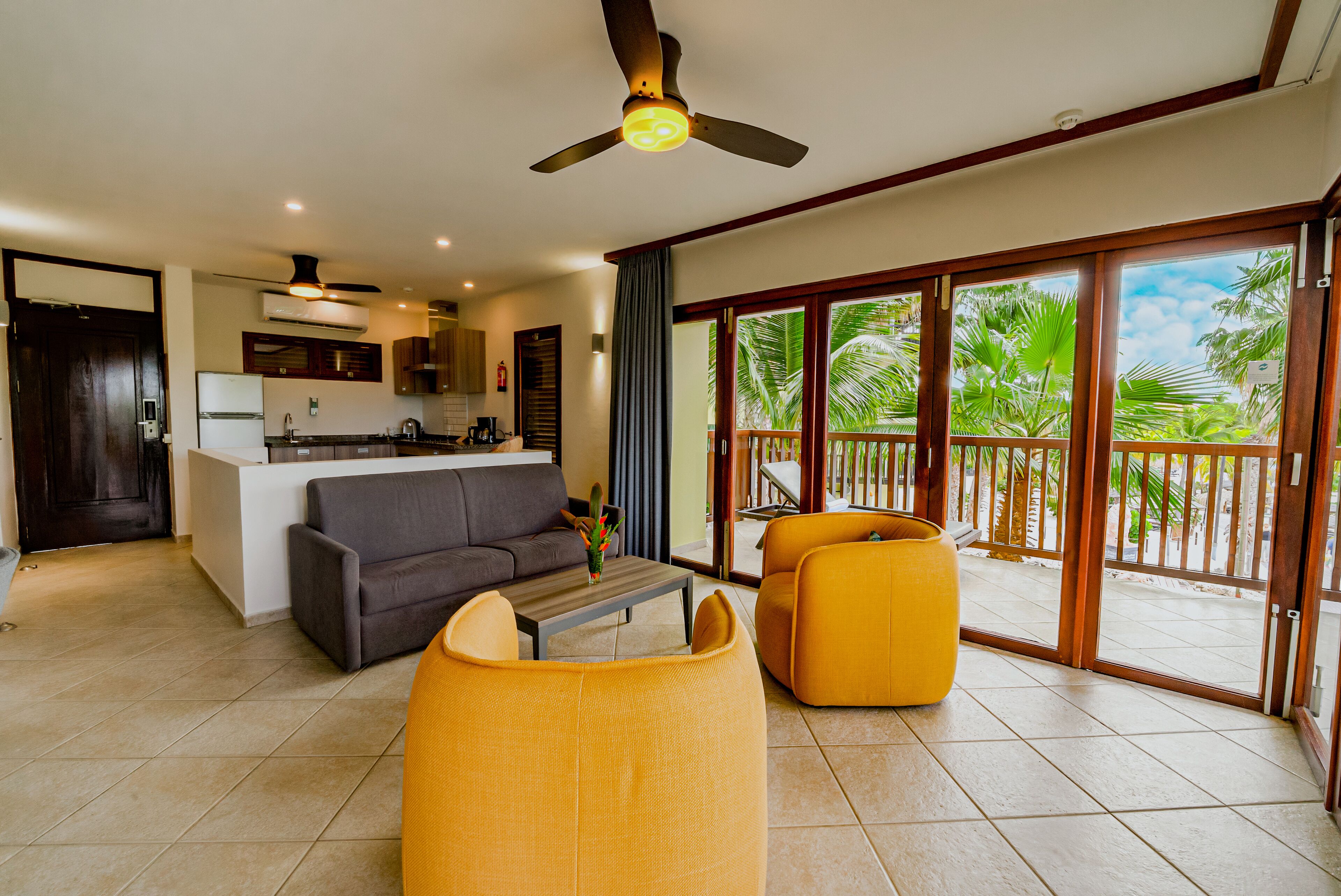 A spacious living area with a sofa, armchairs, and a coffee table. A kitchen is visible in the background, and there is a balcony with a view of palm trees.