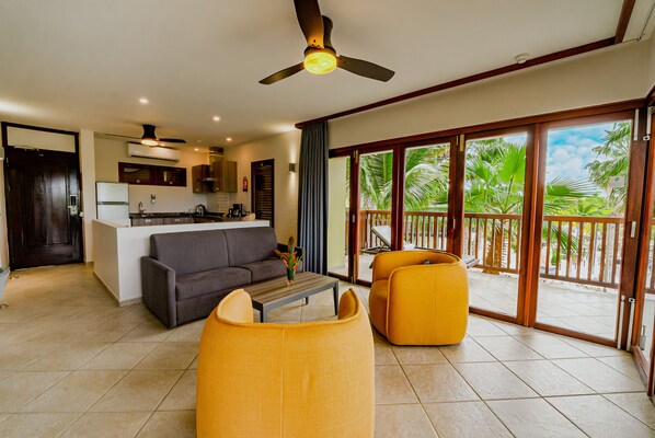 Amplia sala de estar con un sofá, sillones y una mesa de centro. Se ve una cocina al fondo, y un balcón con vistas a palmeras.