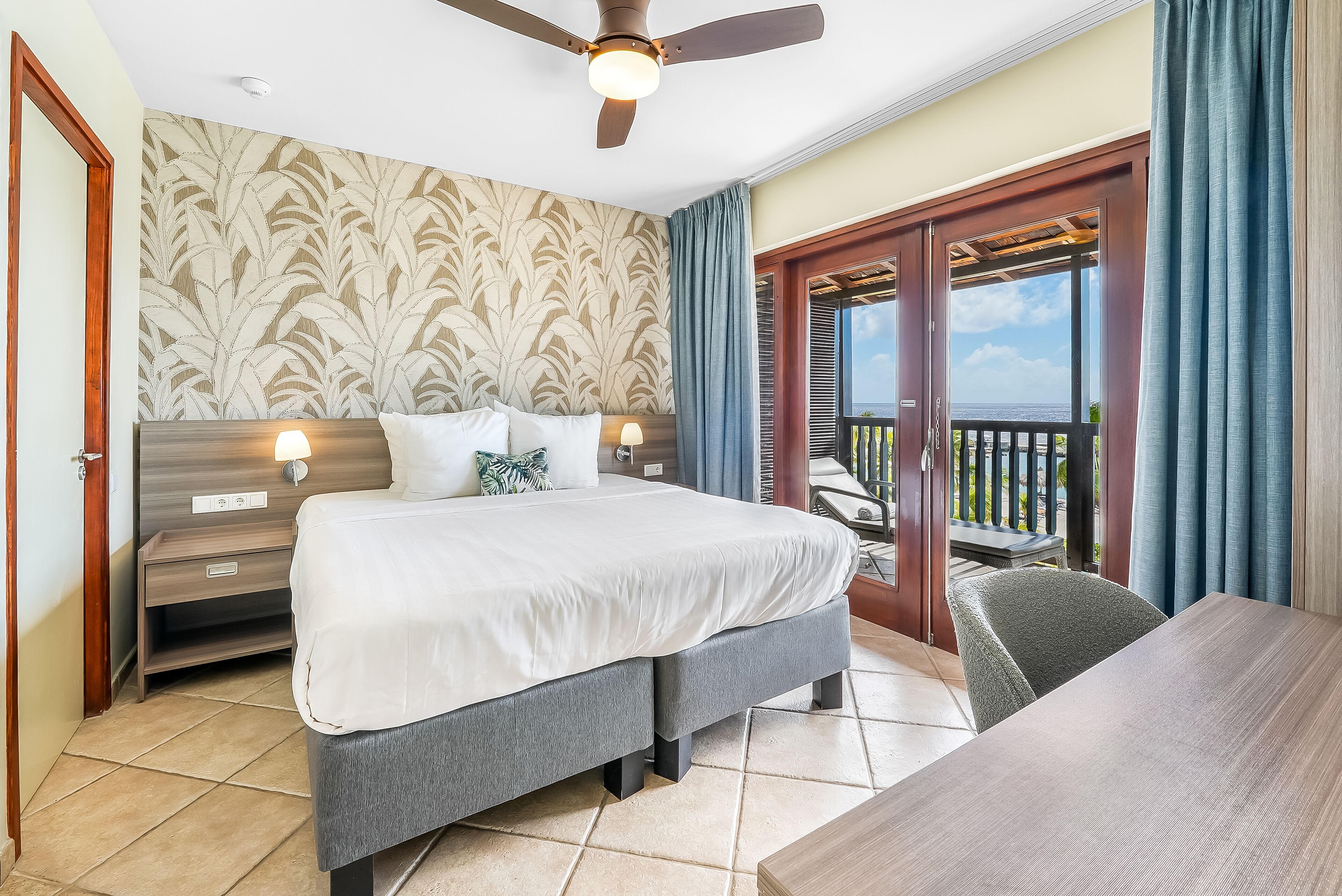 A bedroom with a bed, bedside tables, a desk, and a balcony with a view.