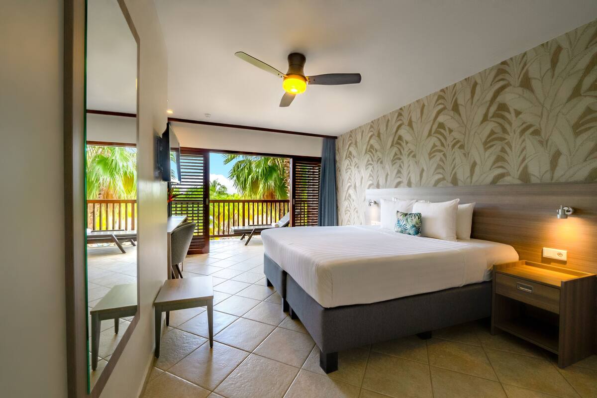 A hotel room with a bed, bedside table, and a view of a balcony with outdoor furniture.