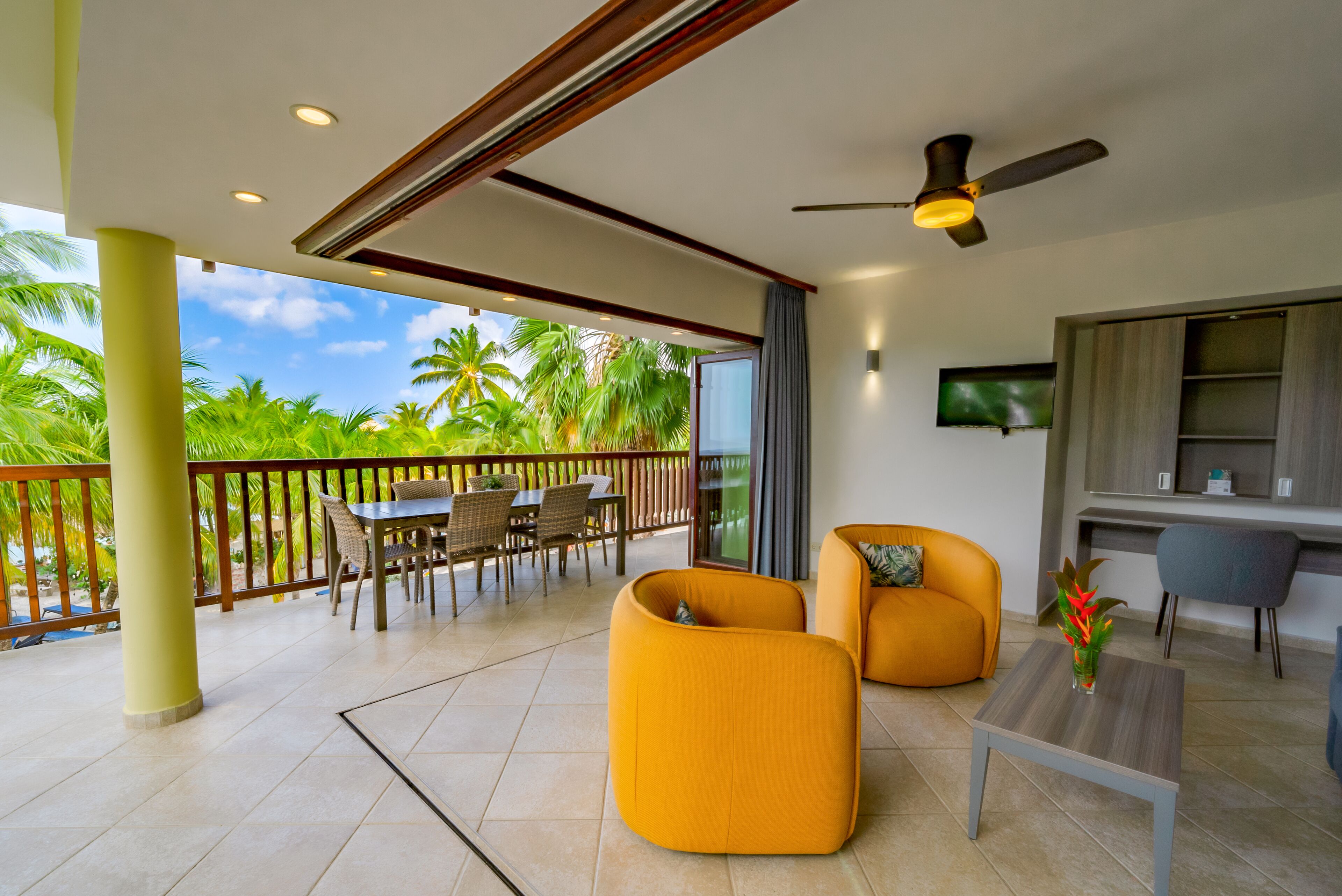 A modern outdoor living area with a dining set, two orange sofas, a small table, and a ceiling fan.
