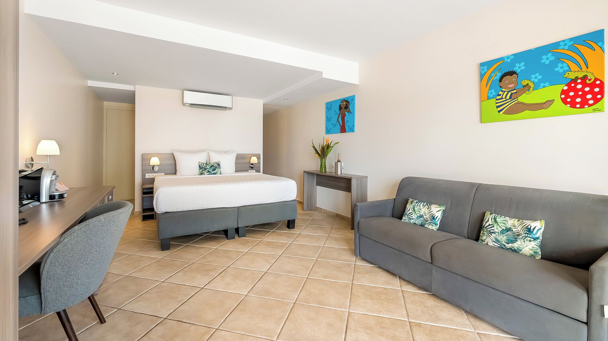 A hotel room with a bed, a grey sofa, a desk with a chair, and two wall art pieces.