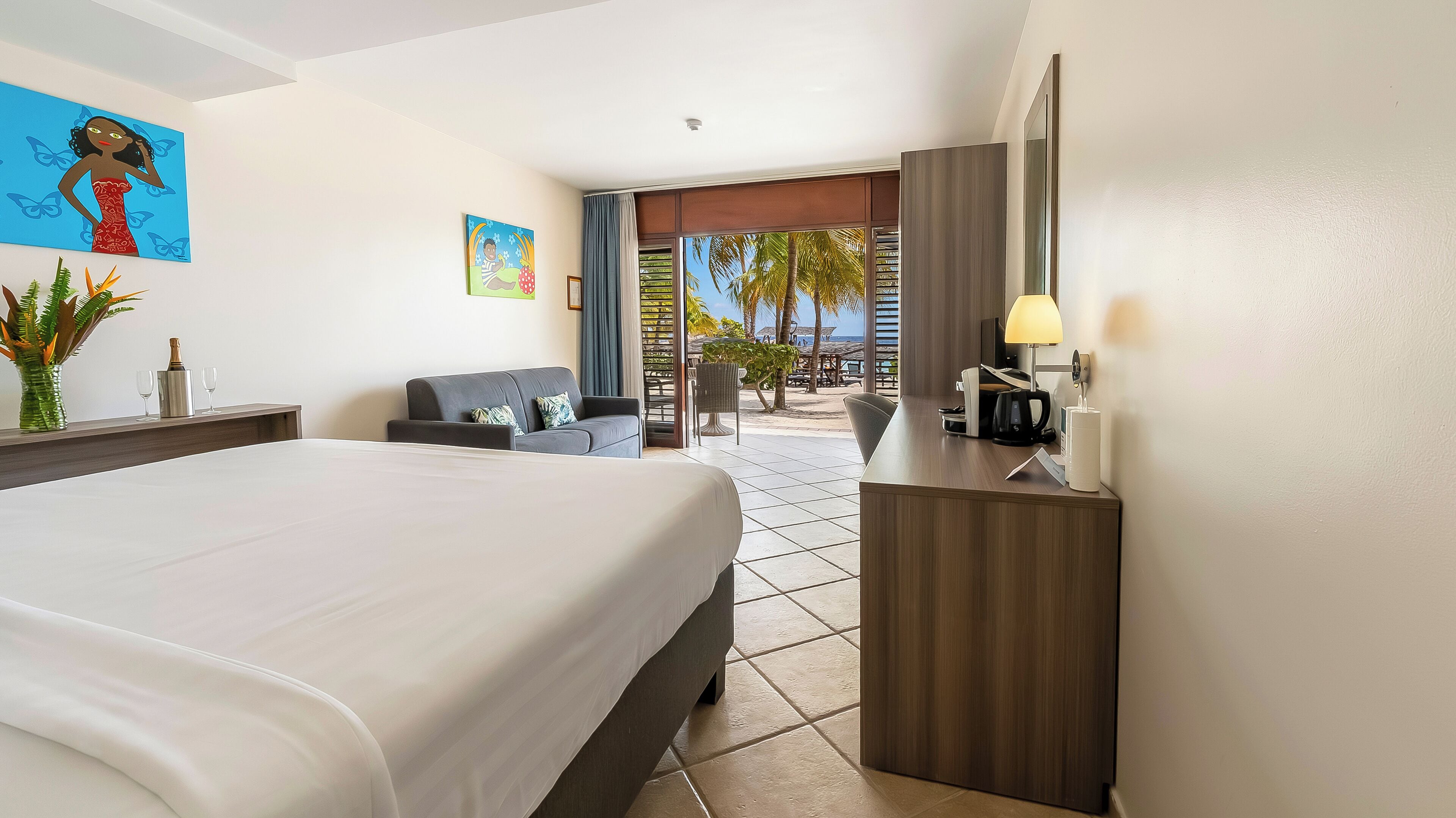 Une chambre d’hôtel avec un grand lit, un bureau avec une lampe, une chaise, une petite table avec une bouteille et des verres, et une vue sur la plage et les palmiers.