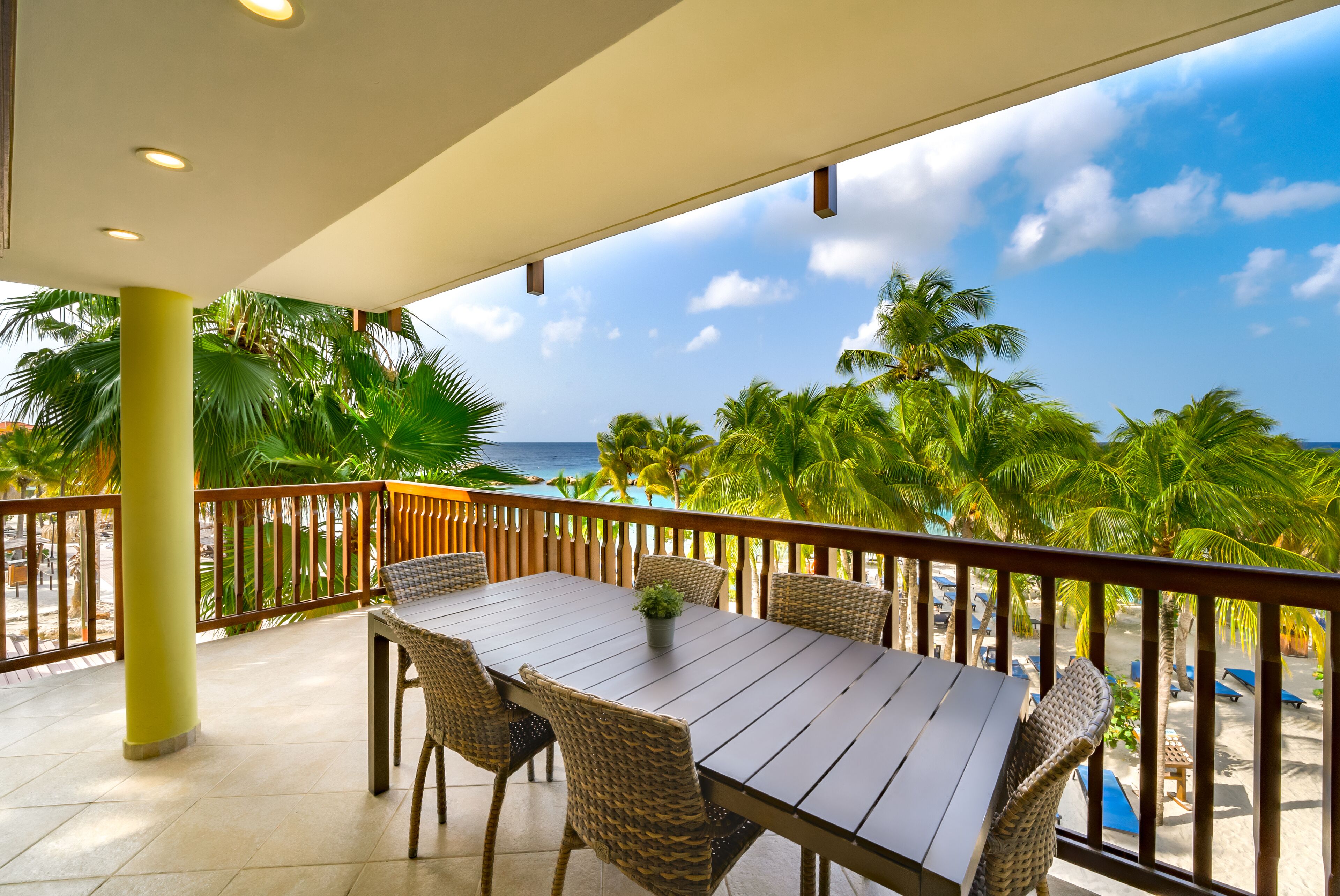 A wooden table with wicker chairs on a balcony overlooking a beach with palm trees and the ocean.