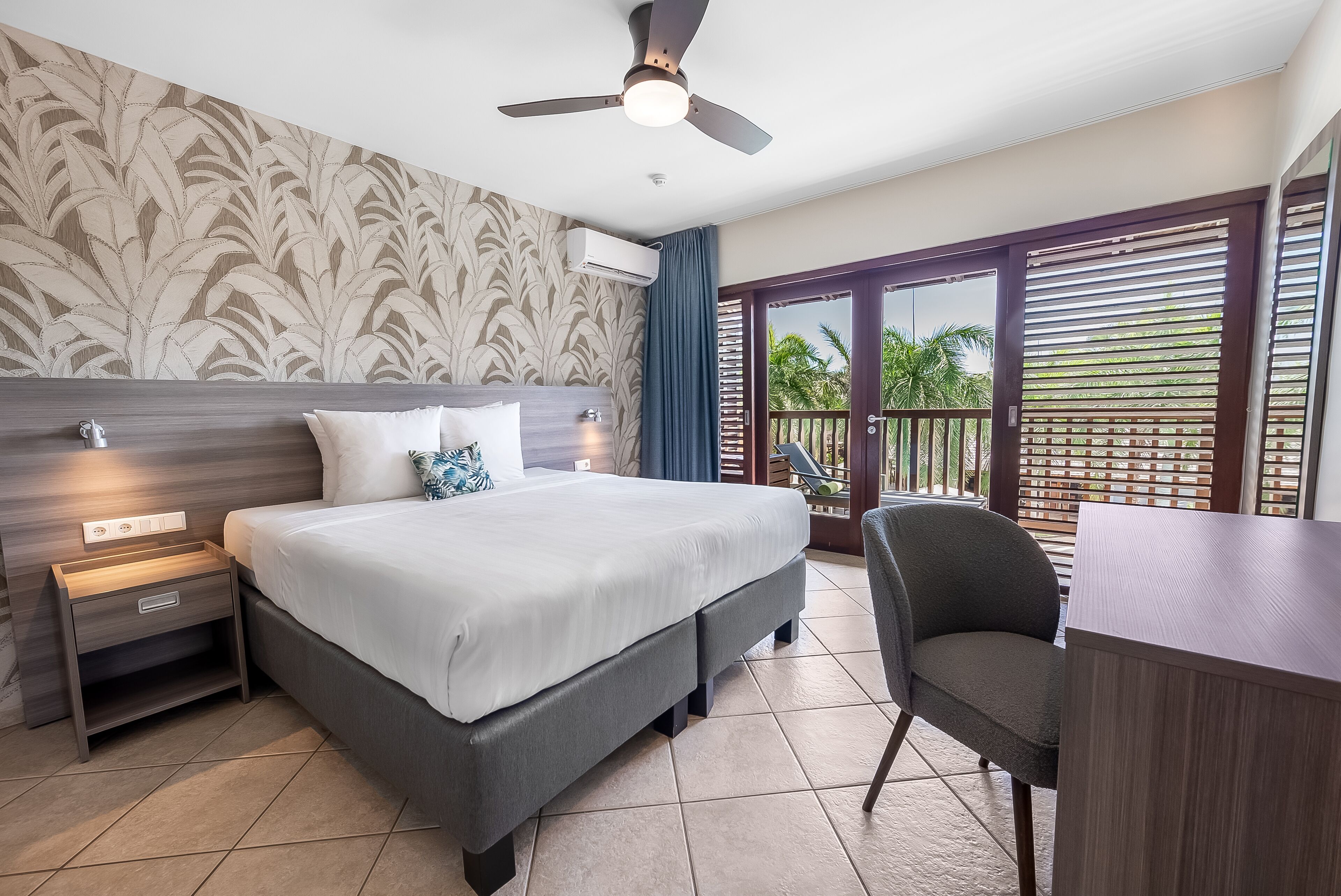 Une chambre d’hôtel avec un grand lit, un bureau, une chaise, un ventilateur de plafond et un balcon offrant une vue sur les palmiers.