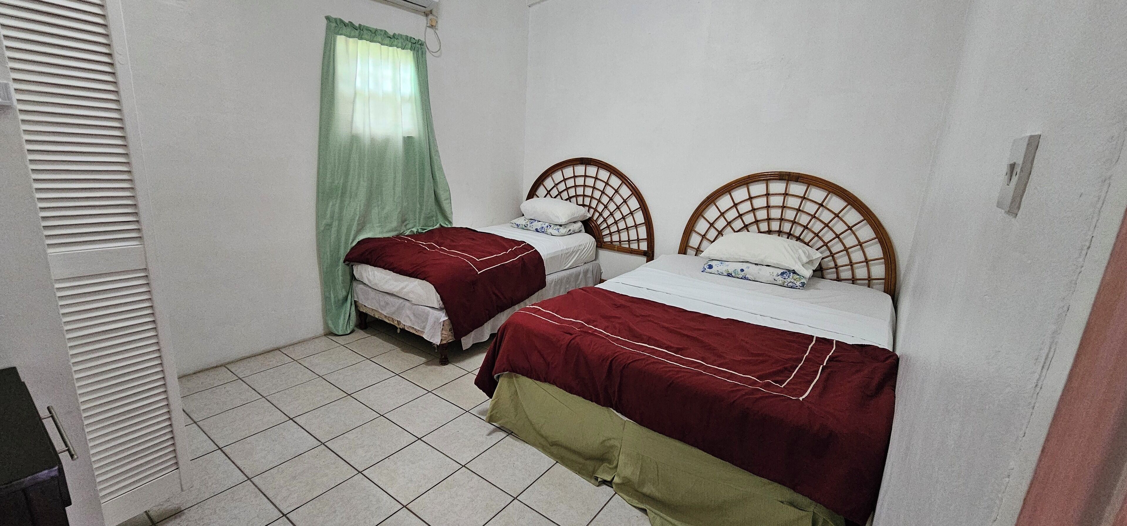 Una habitación pequeña y limpia con dos camas individuales, una ventana con cortina verde y suelo de baldosas.