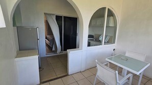 Un pequeño balcón cerrado con una mesa, sillas, un refrigerador y una ventana que da al interior.