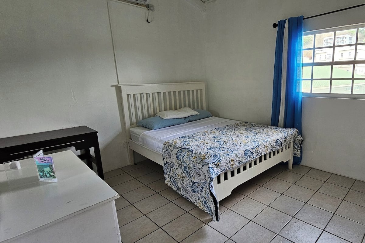 A small, clean room with a single bed, a window with blue curtains, and a tiled floor.
