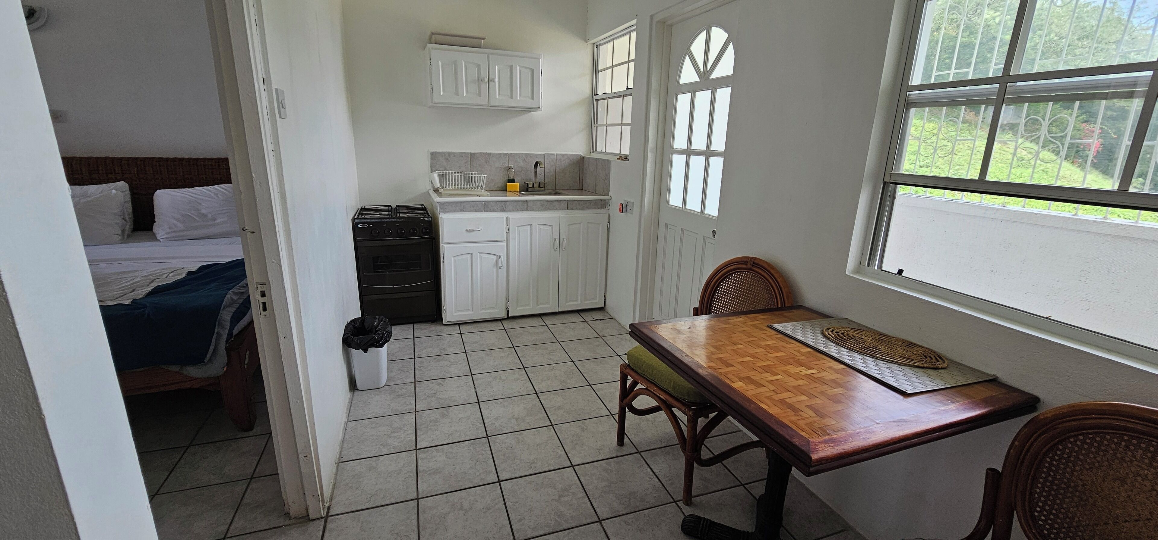 Een kleine kamer met een bed, een kitchenette, een eettafel en een raam met uitzicht op groen.