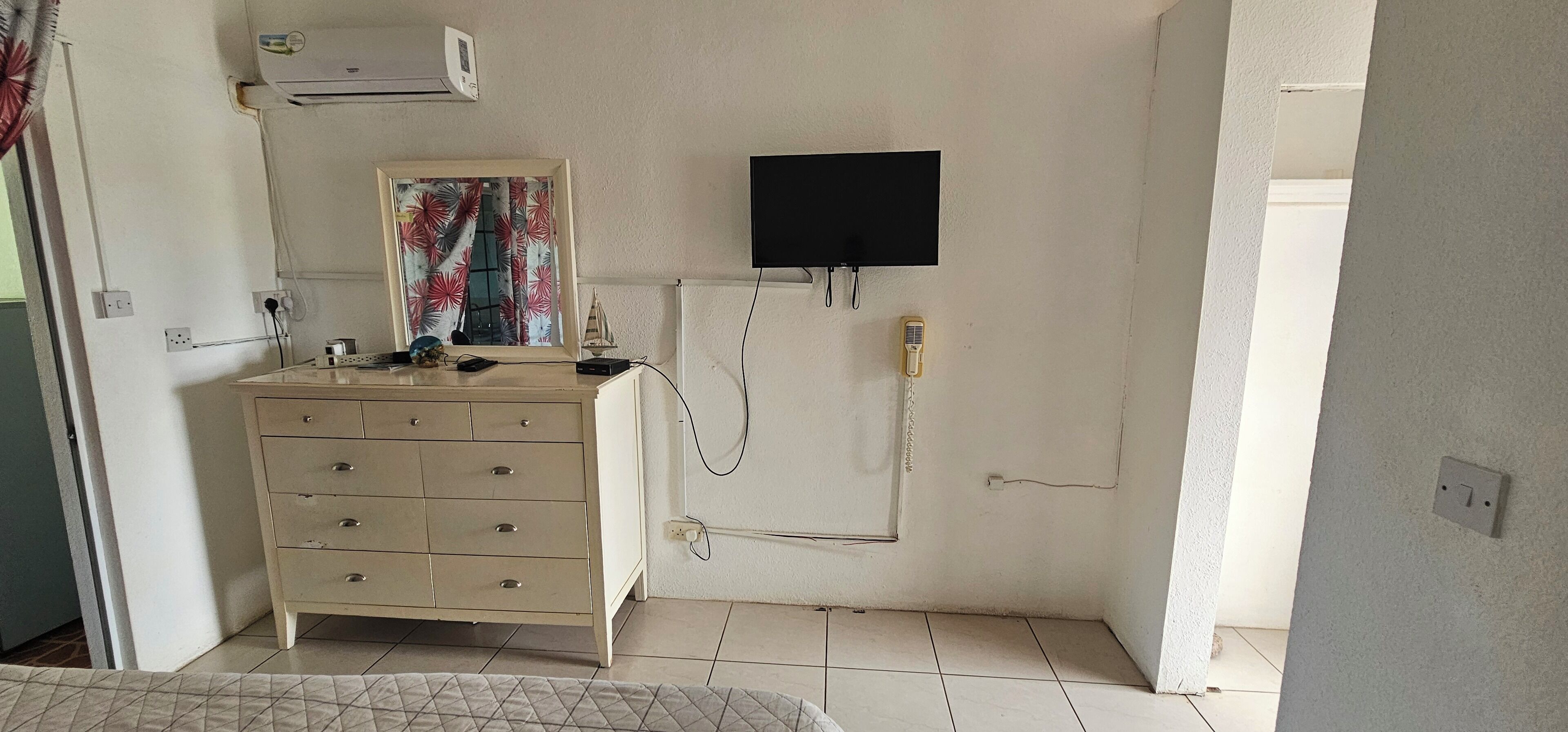 A small, well-lit room with a beige dresser, a flat-screen TV mounted on the wall, and a telephone.