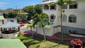 Un edificio de varios pisos con techo verde, un espacio de estar al aire libre cubierto y un césped bien cuidado con palmeras.