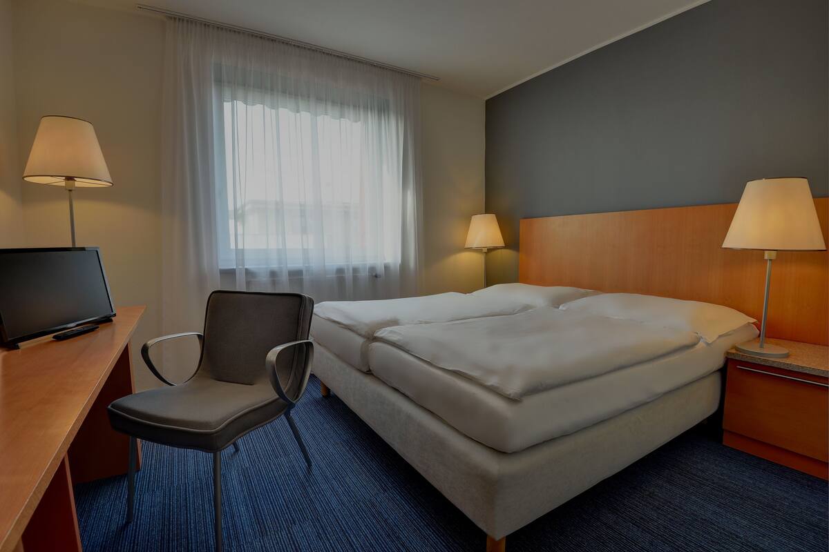 A hotel room with a bed, a desk with a television, two chairs, and two bedside lamps.