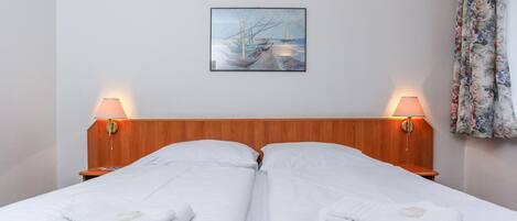 A neatly made double bed with white linens, two bedside lamps, and a painting on the wall.