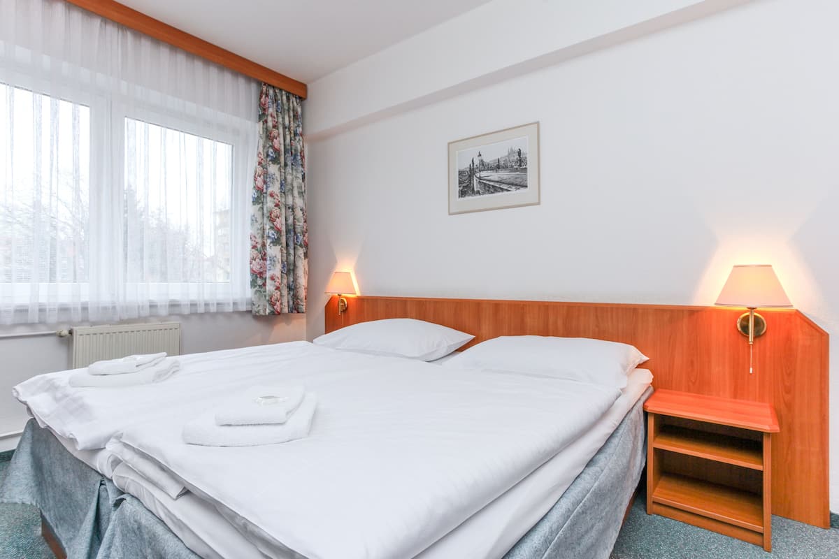 Quarto de hotel com cama bem arrumada, duas luminárias de cabeceira, um quadro na parede e uma janela com cortinas translúcidas.