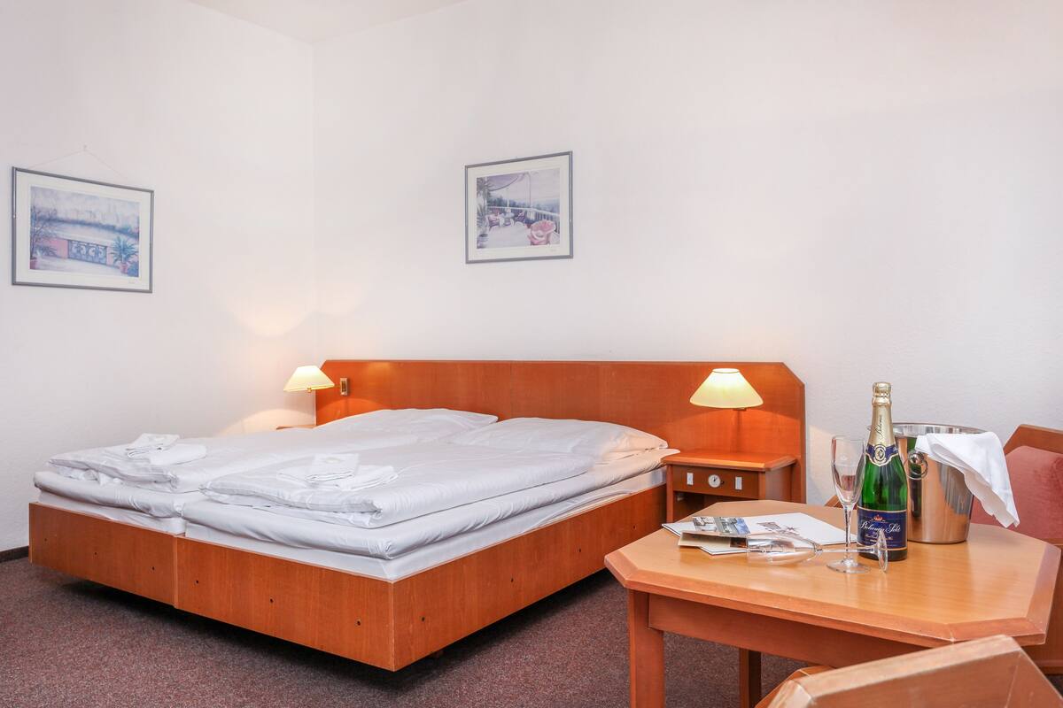 Cama bem arrumada com cabeceira de madeira, duas luminárias de apoio, uma mesa com champanhe, taças e um menu.