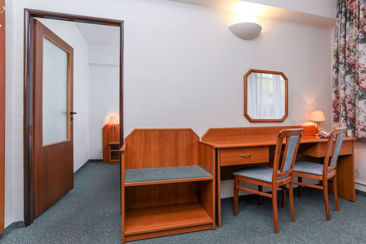 Quarto compacto com escrivaninha de madeira, duas cadeiras, um espelho e uma pequena mesa de cabeceira com luminária.