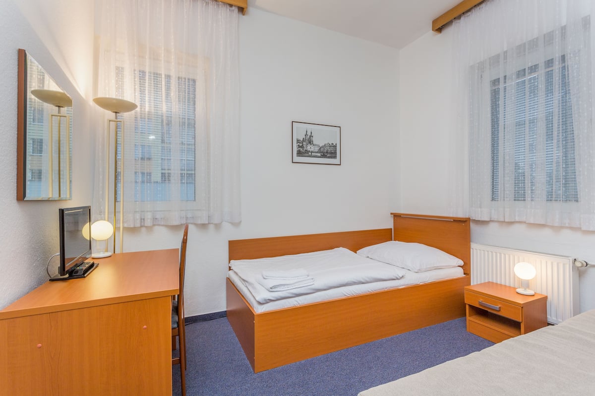 Quarto de hotel com cama de madeira, mesa de cabeceira com luminária, escrivaninha com computador e luminária, e janela com cortinas finas.