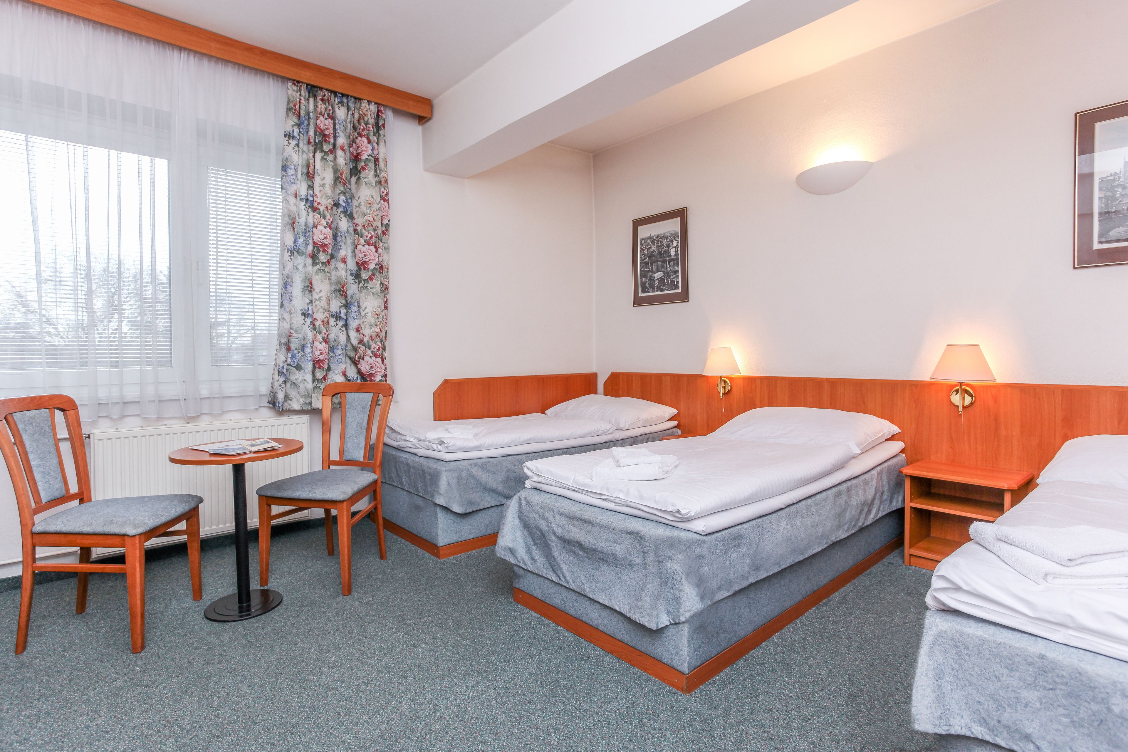 Hotellihuone, jossa on kaksi erillistä sänkyä, pieni pöytä tuoleineen, ikkuna verhoineen ja seinään kiinnitetty valaisin.