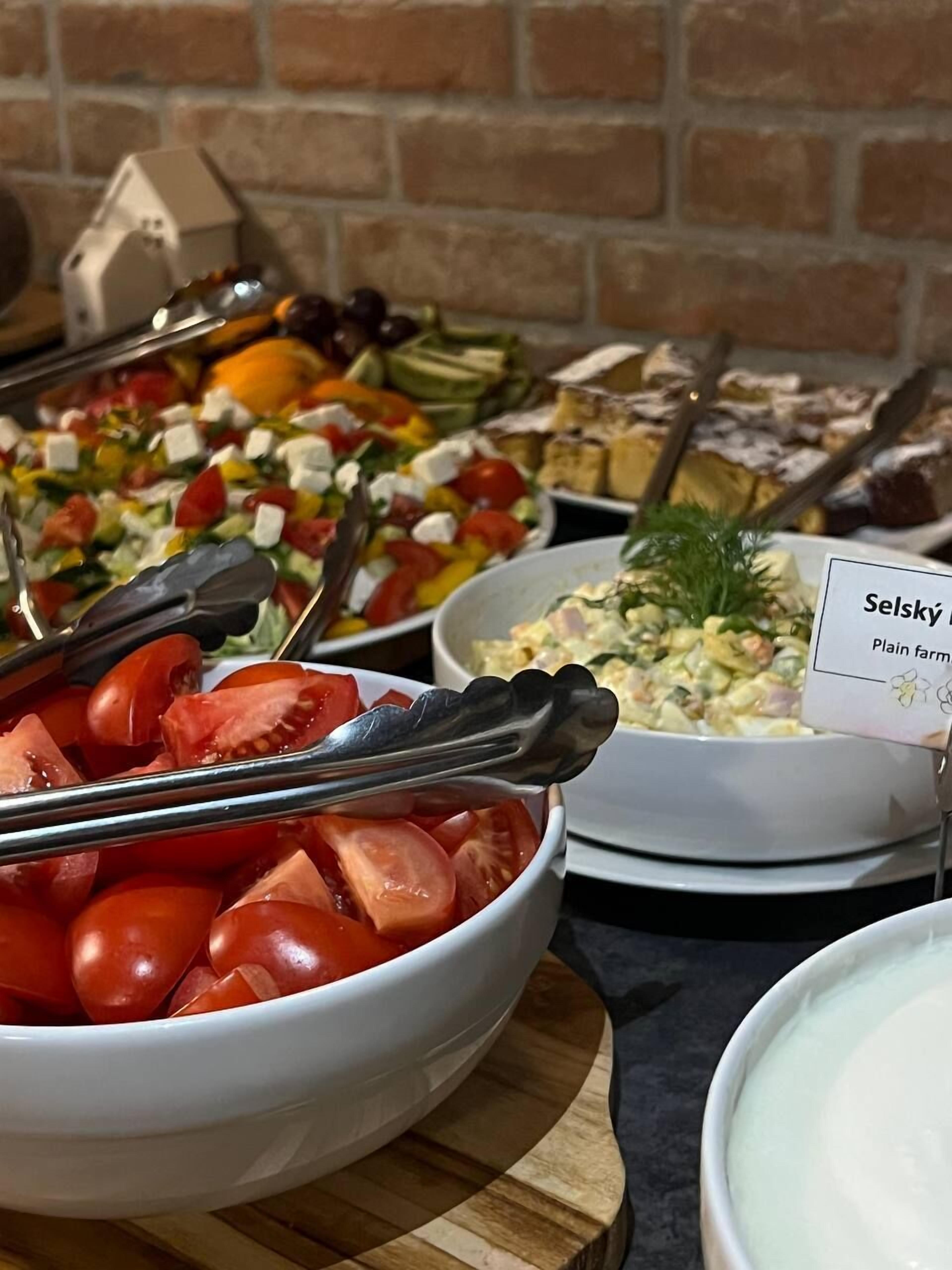 Buffet amb diferents plats, incloent un bol de tomàquets tallats, una ensalada i un cartell que diu 'Selský Plain Farm'.