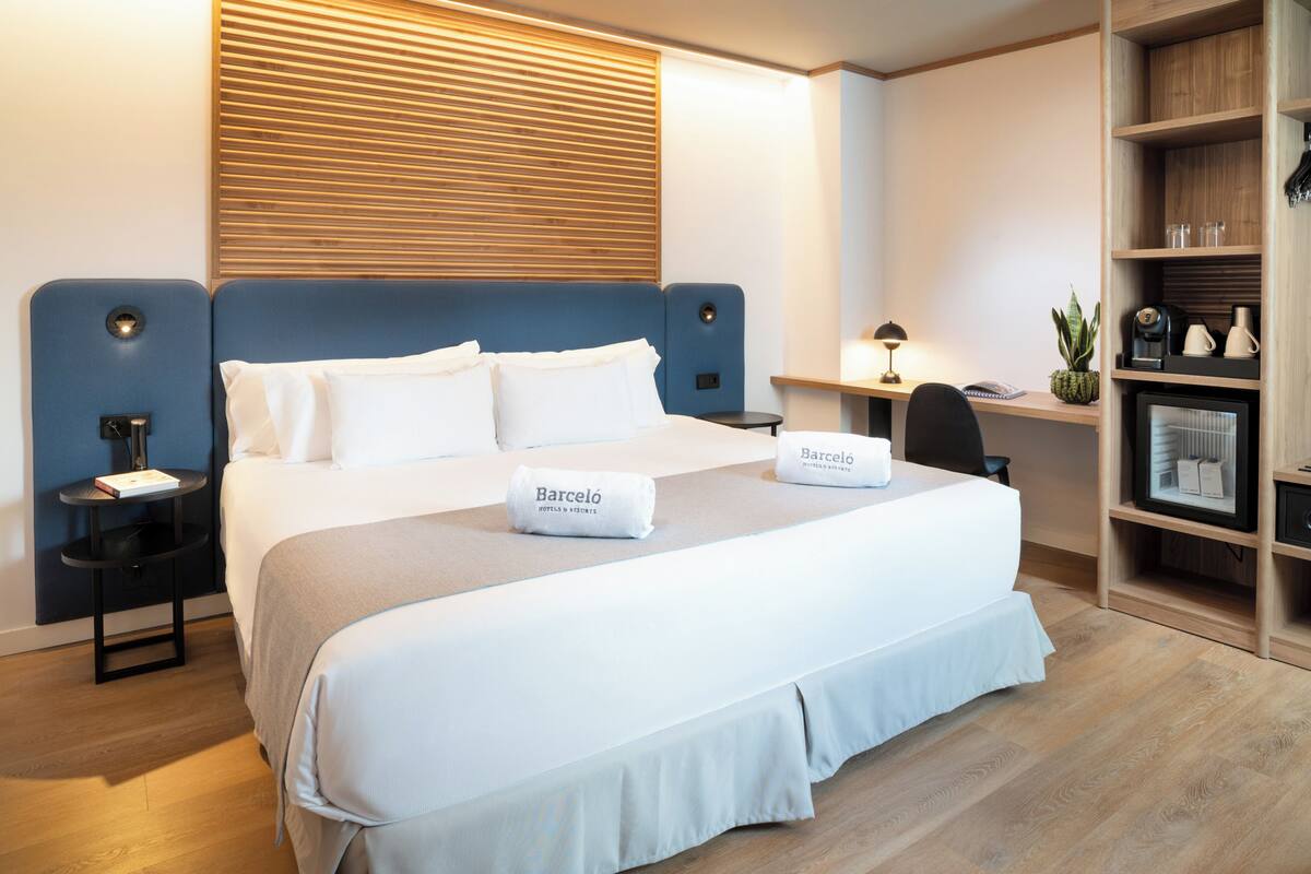 Habitación de hotel con una cama grande, una mesita de noche con lámpara, una pequeña mesa auxiliar, un estante de madera con objetos y una ventana con persianas.