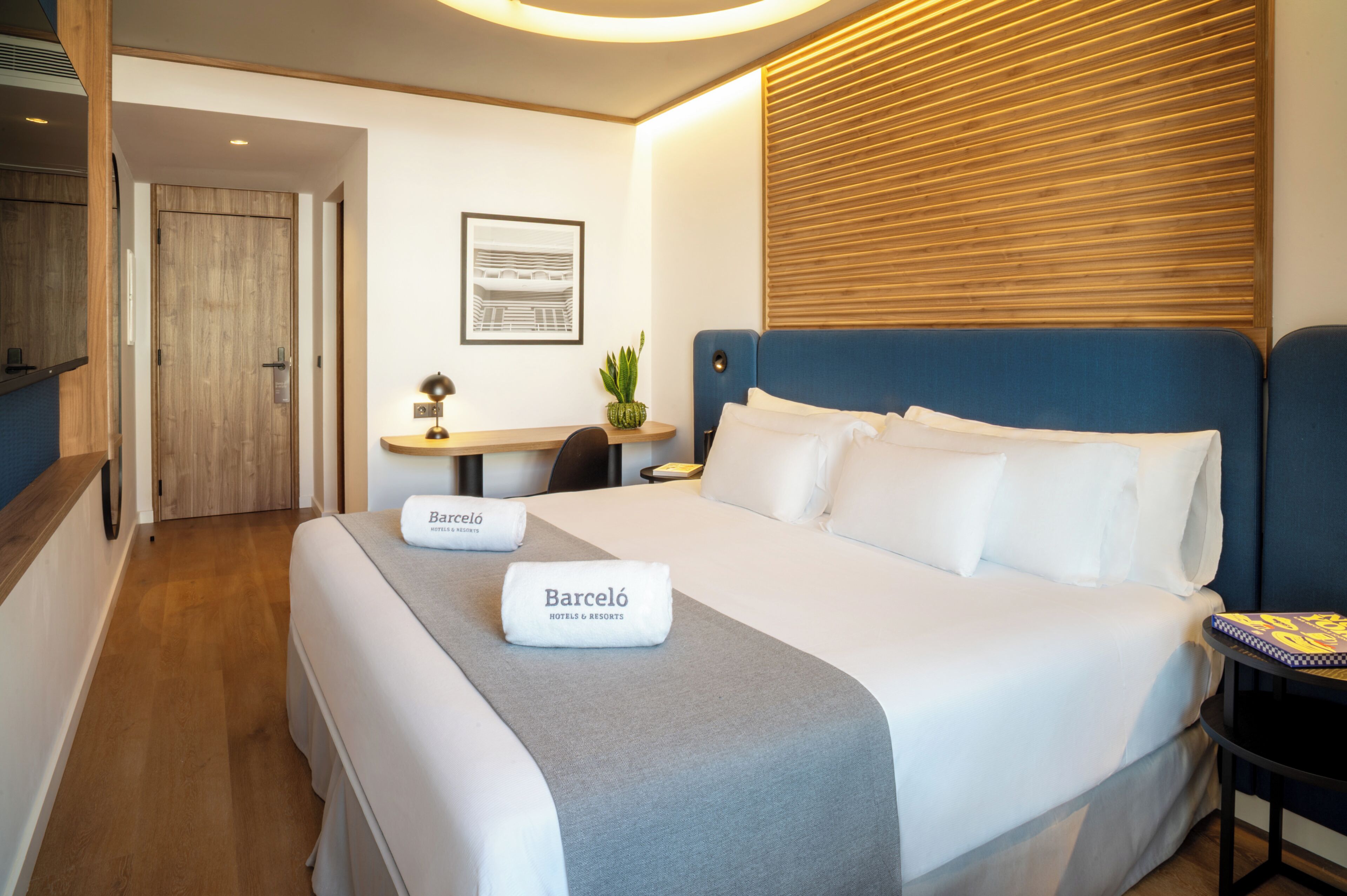 A hotel room with a large bed, a desk, a chair, a small table with a plant, and a wall-mounted shelf.