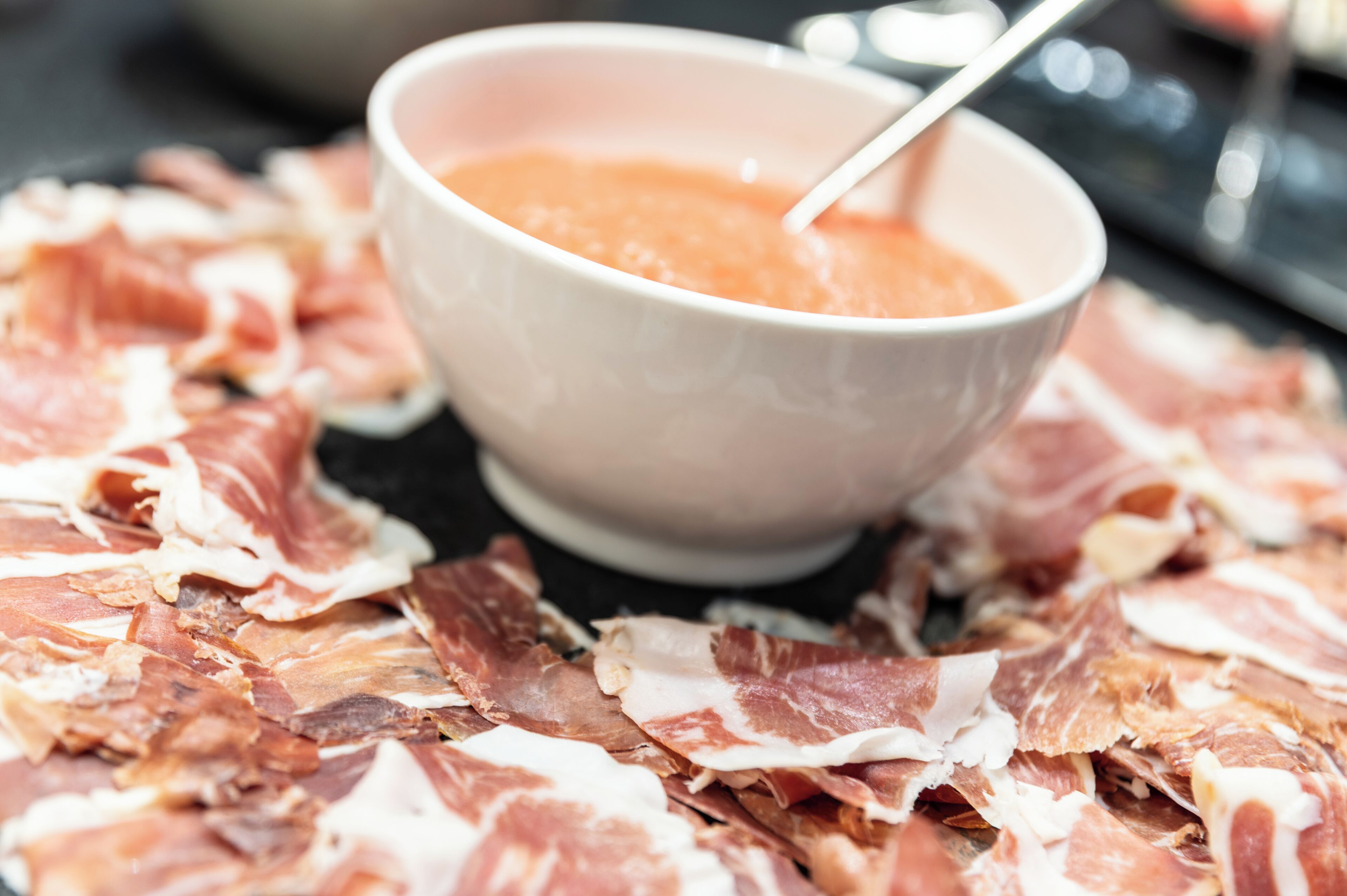 A bowl of sauce with a spoon, surrounded by scattered meat slices.