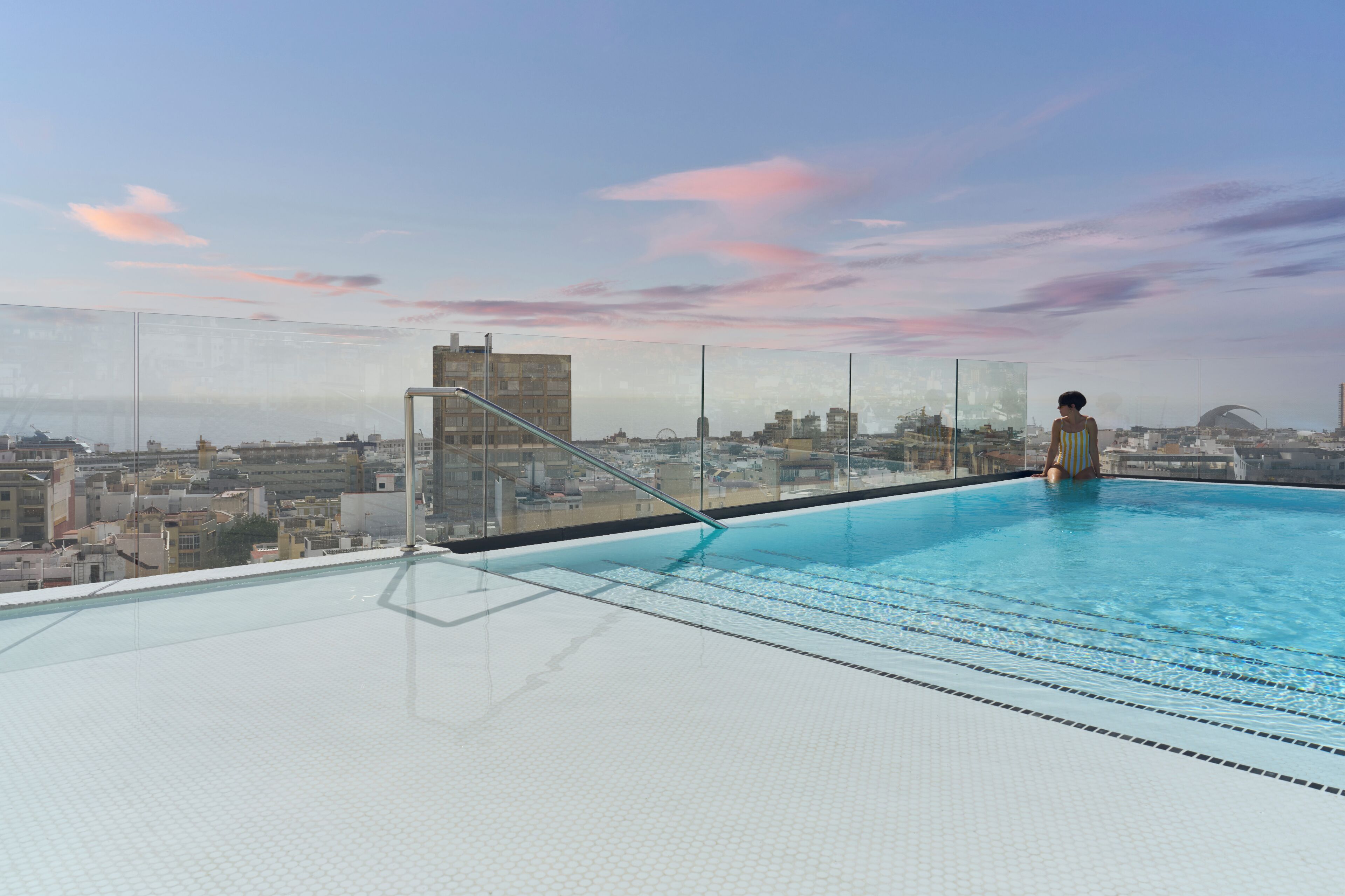 Una piscina infinita en la azotea con vistas a la ciudad, barandilla de cristal y cielo despejado.