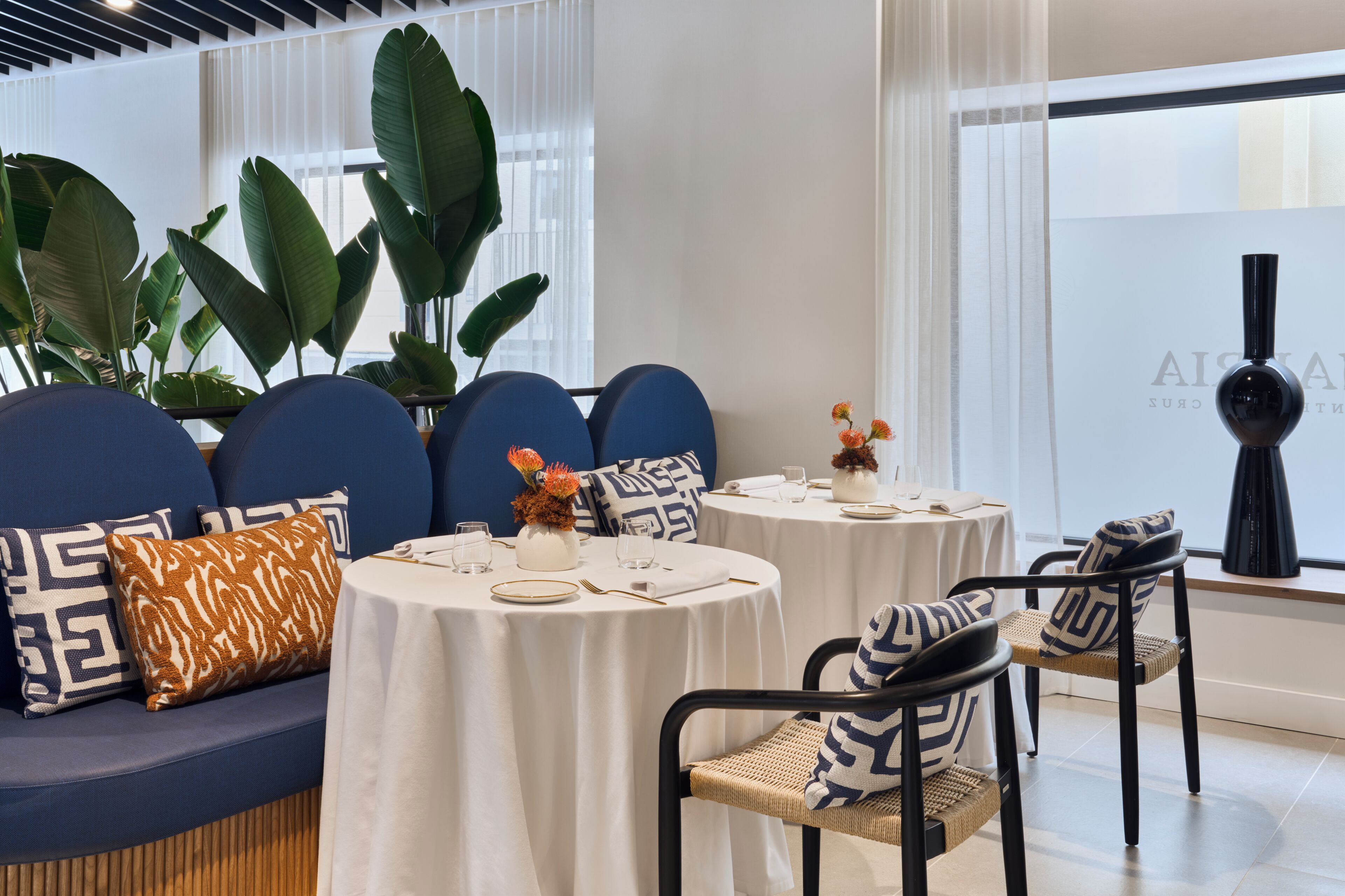 A modern dining area with round tables, blue upholstered seating, and large green plants.