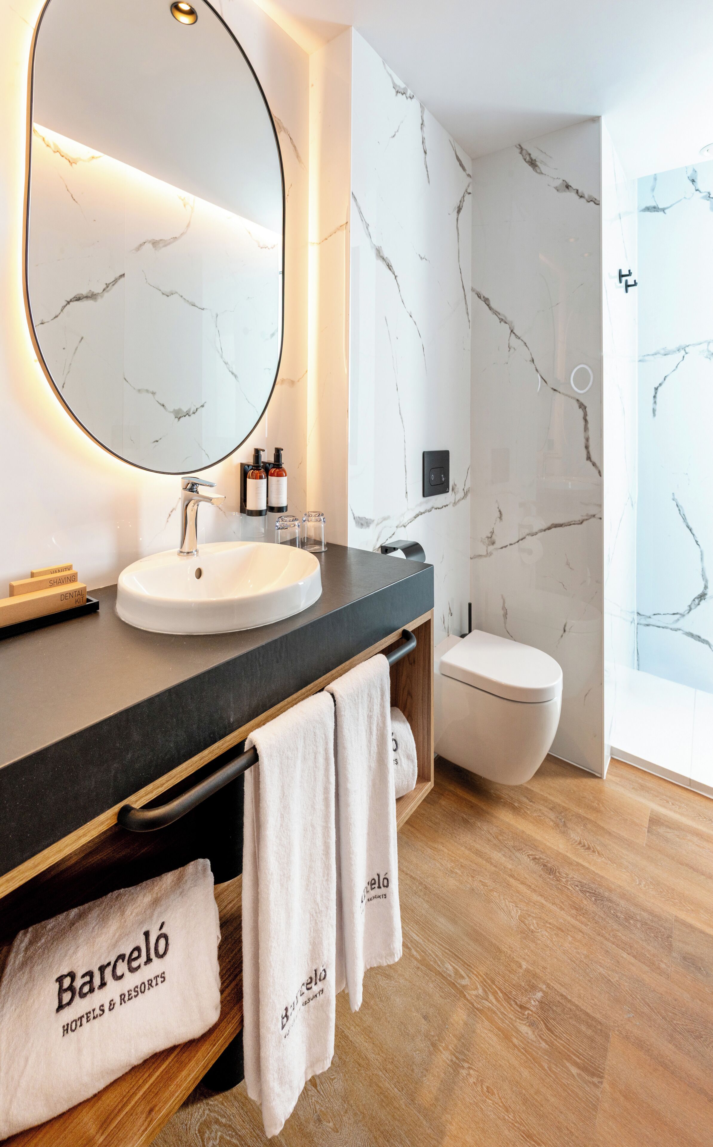 A modern bathroom with a marble backsplash, a round sink, and a toilet. There are towels hanging on a rack and a mirror above the sink.
