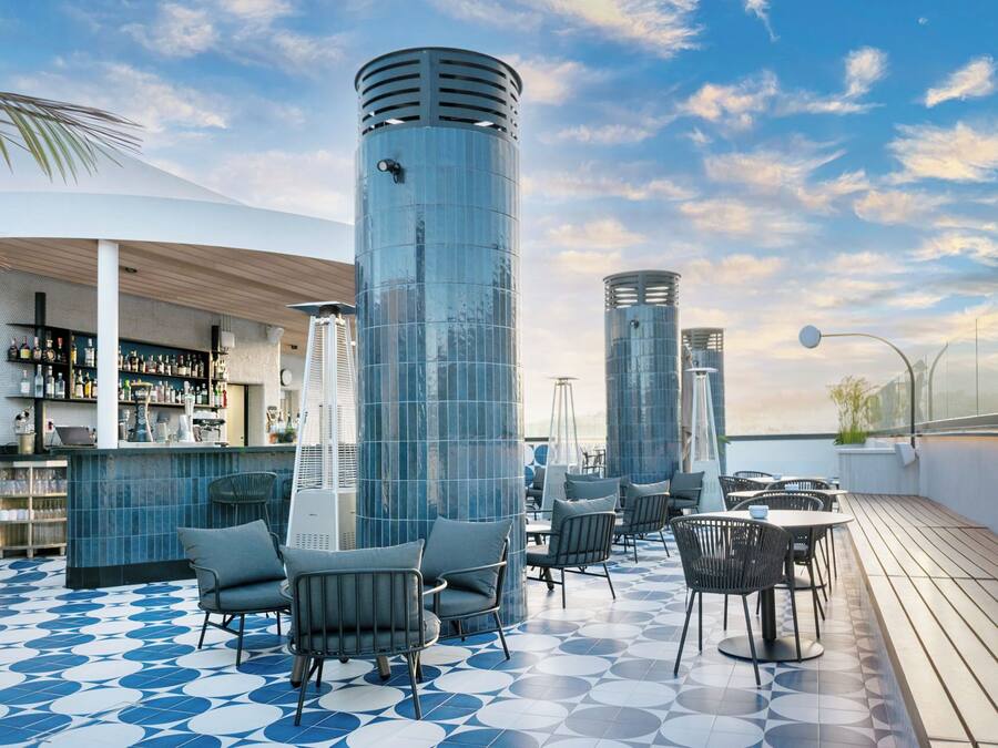 A rooftop bar with a blue and white tiled floor, modern seating, and a bar area with shelves of bottles.