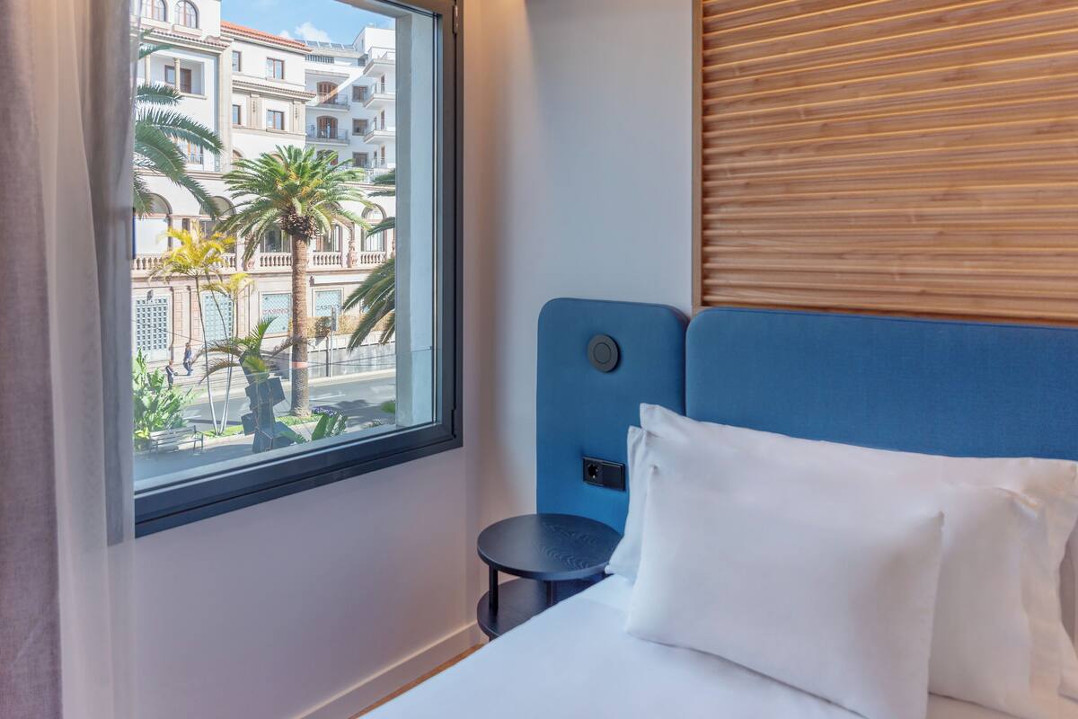 Habitación de hotel con cama, ventana con vistas a edificios, una mesita redonda y cabecera azul.