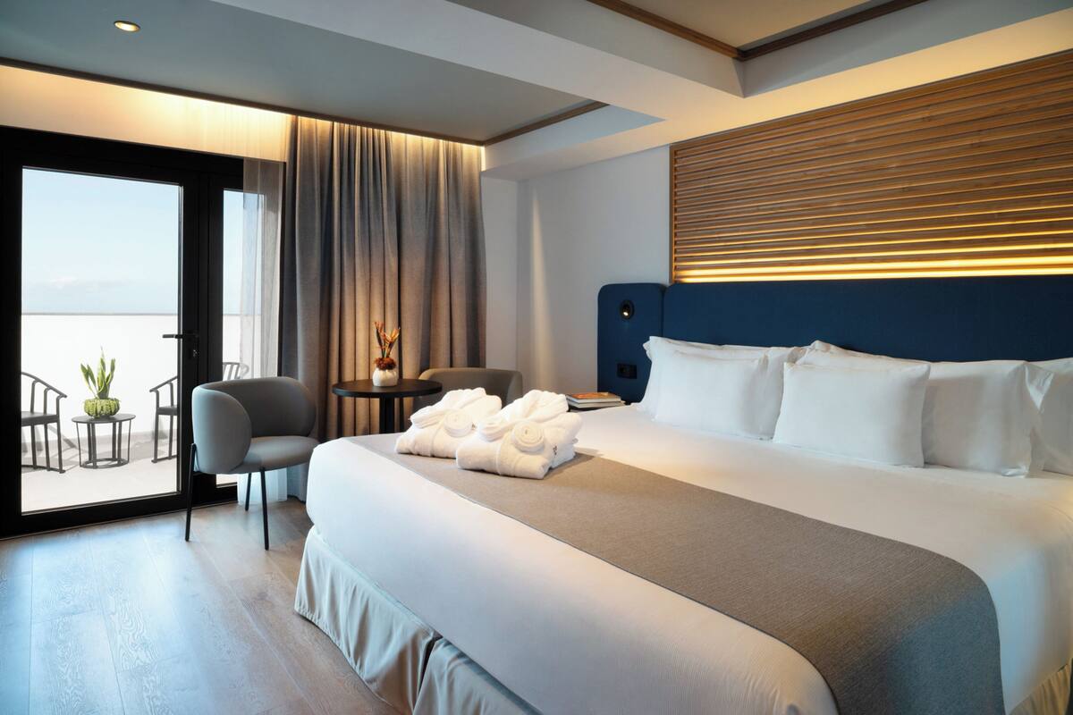 Una habitación de hotel moderna con una cama grande, un área de descanso con sillas, una pequeña mesa con una planta y vistas a través de un ventanal.