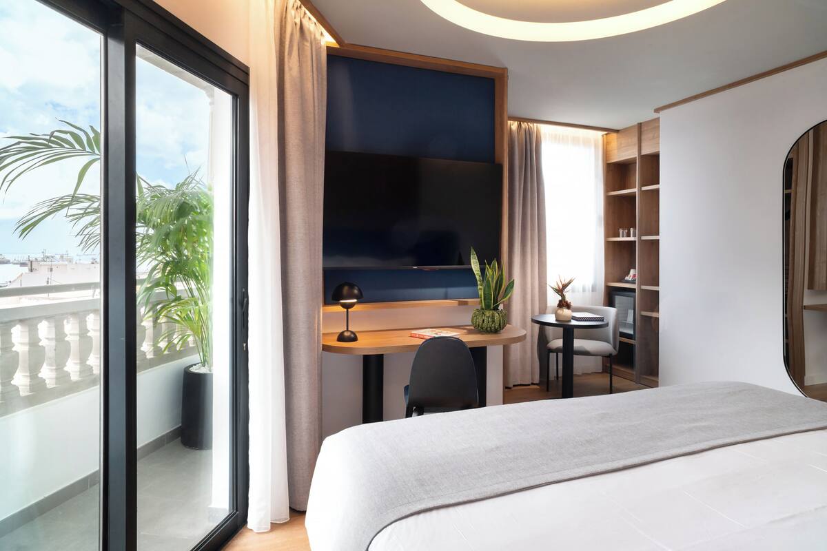 Habitación de hotel con cama, escritorio con televisor, una mesita, una silla, planta en maceta y vista a la ciudad.