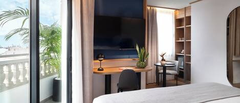 Habitación de hotel con cama, escritorio con televisor, una mesita, una silla, planta en maceta y vista a la ciudad.