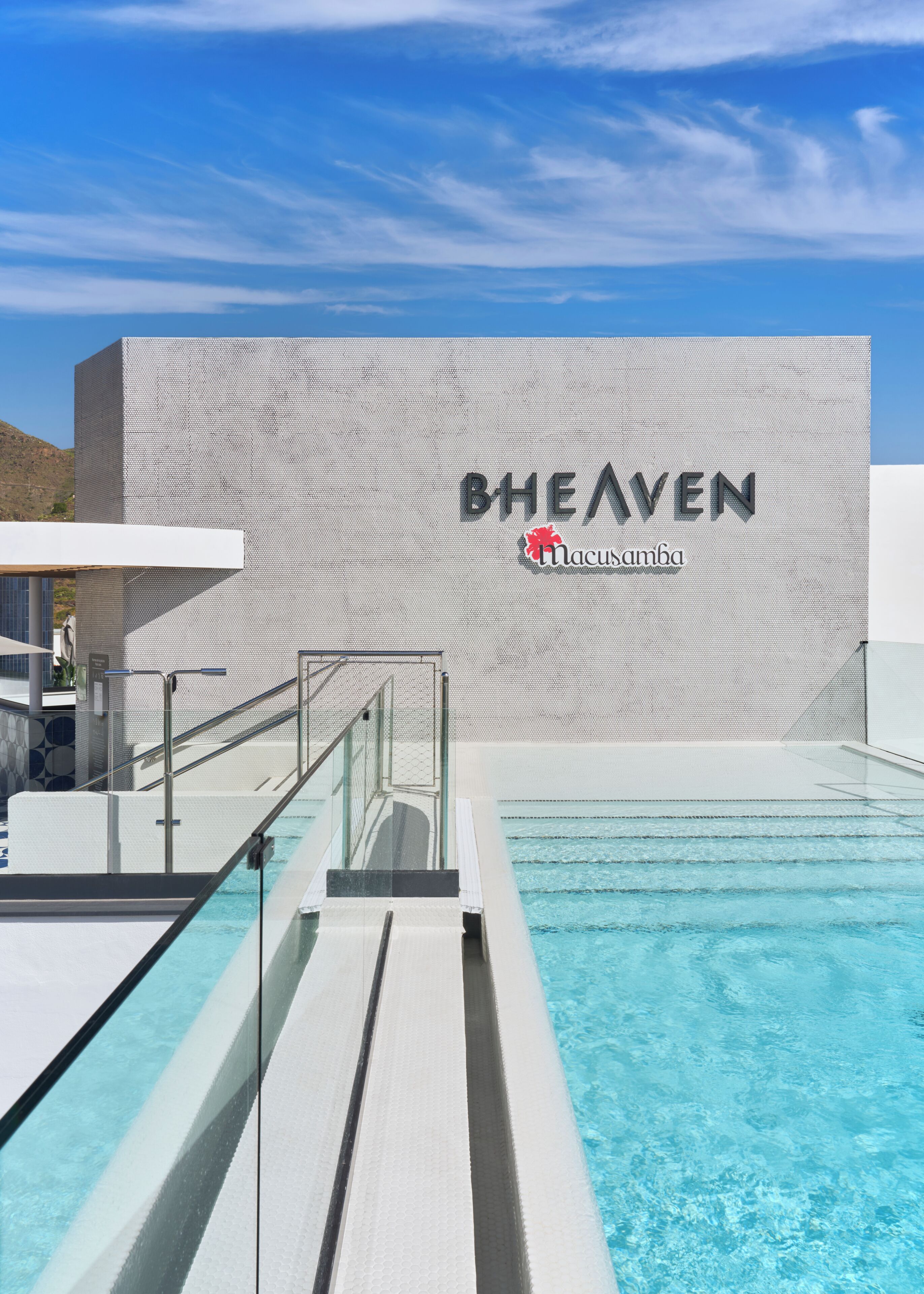 A swimming pool with a modern building in the background, a sign reading 'BHEAVEN Macusanga', and a clear blue sky.