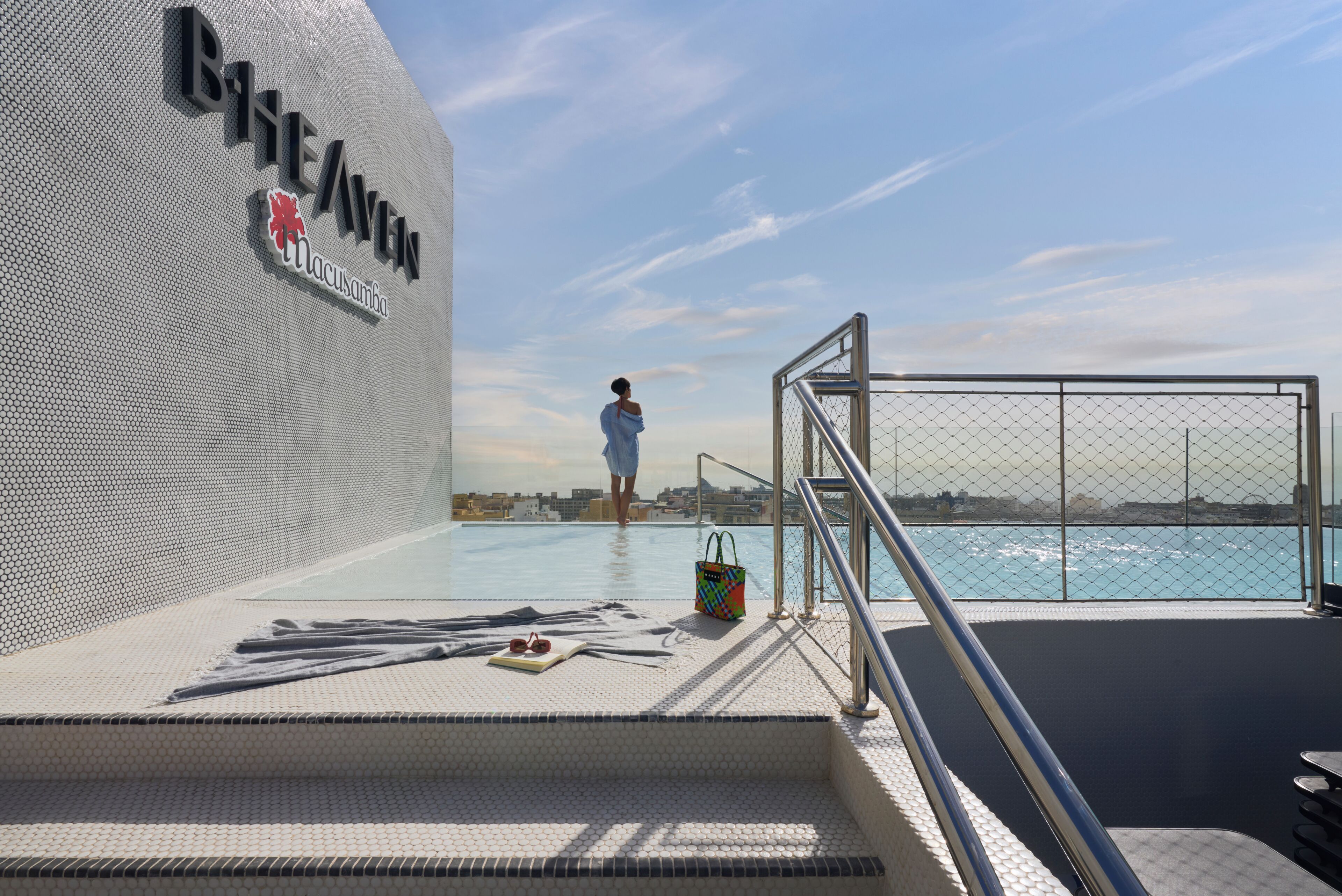 A poolside area with a person walking towards the water, a sign reading 'B1HEAVEN', and a view of the sea.
