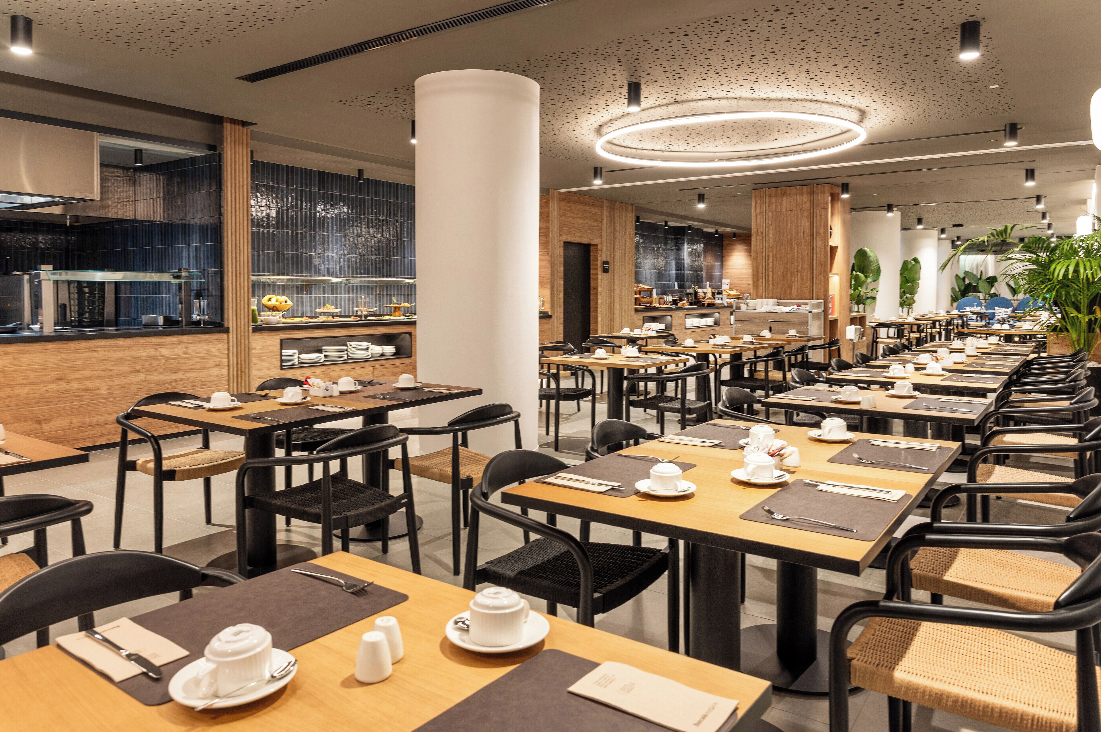 A modern restaurant with multiple dining tables and chairs, a kitchen area, and a decorative ceiling light.